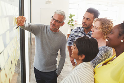Mature businessman and his diverse team brainstorming with yellow adhesive notes on a glass wall in a bright modern office